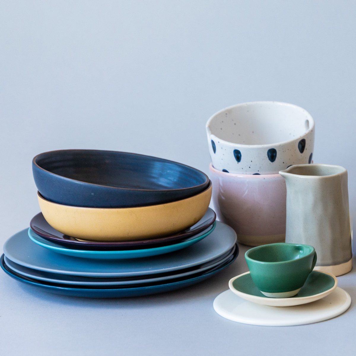 Stoneware on a grey table: an ink bowl on a sun bowl and a stack of plates, a dotted bowl on a blush bowl, a grey jug and a green cup on its saucer