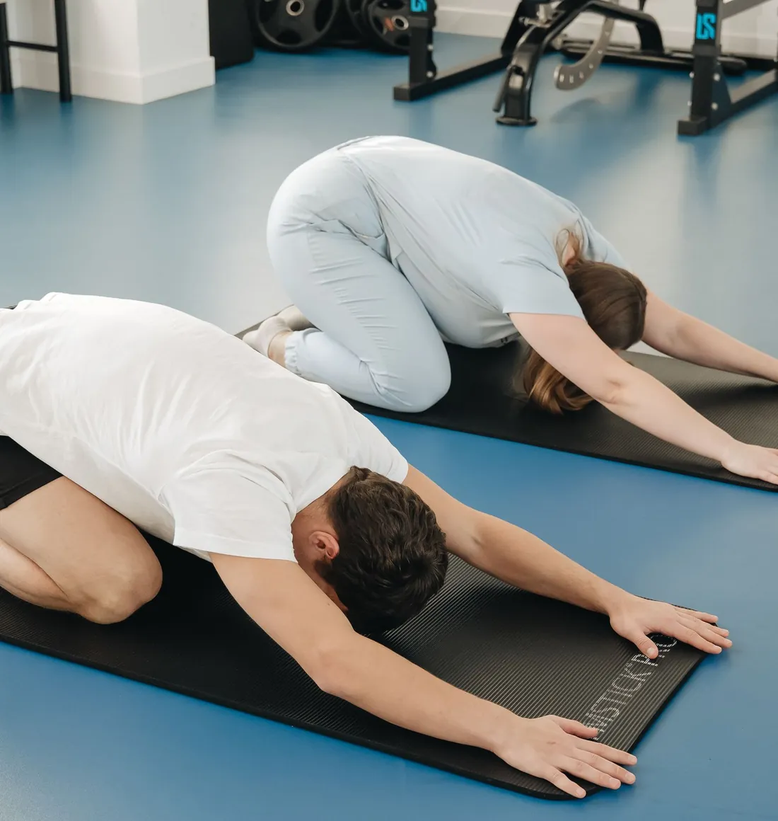 Two people stretching on blue exercise mats in a bright clinic