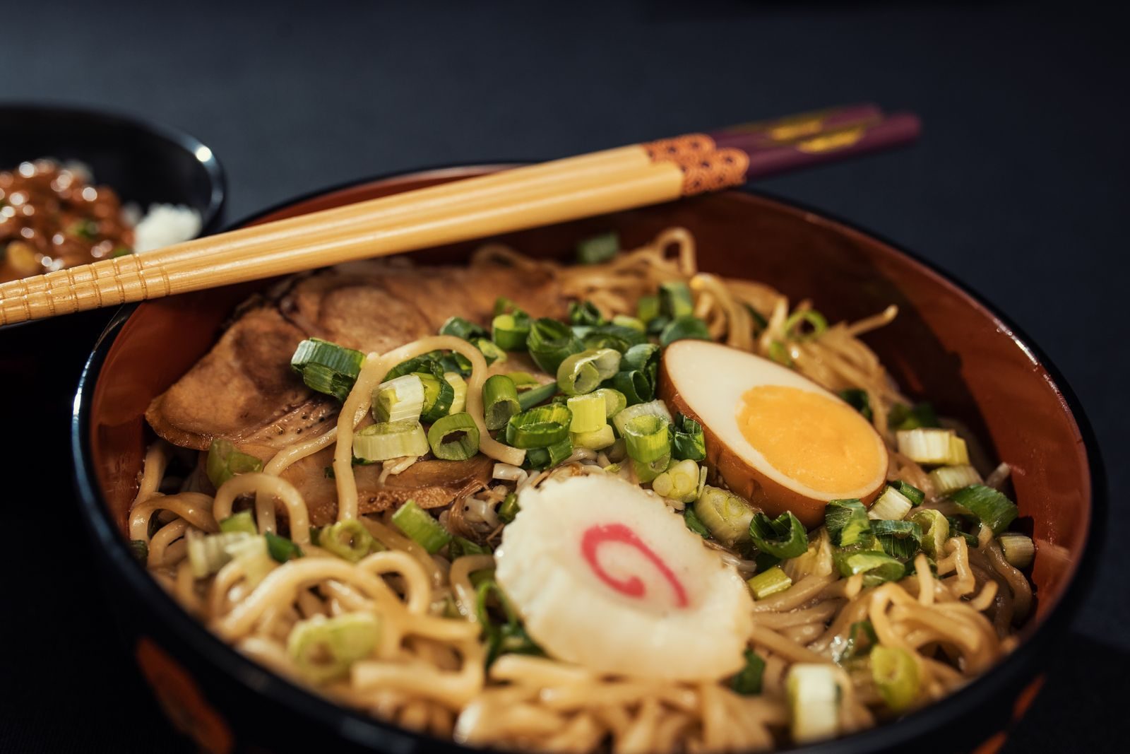 Shoyu ramen with a marinated egg, fish cake, pork and scallions in a dark bowl
